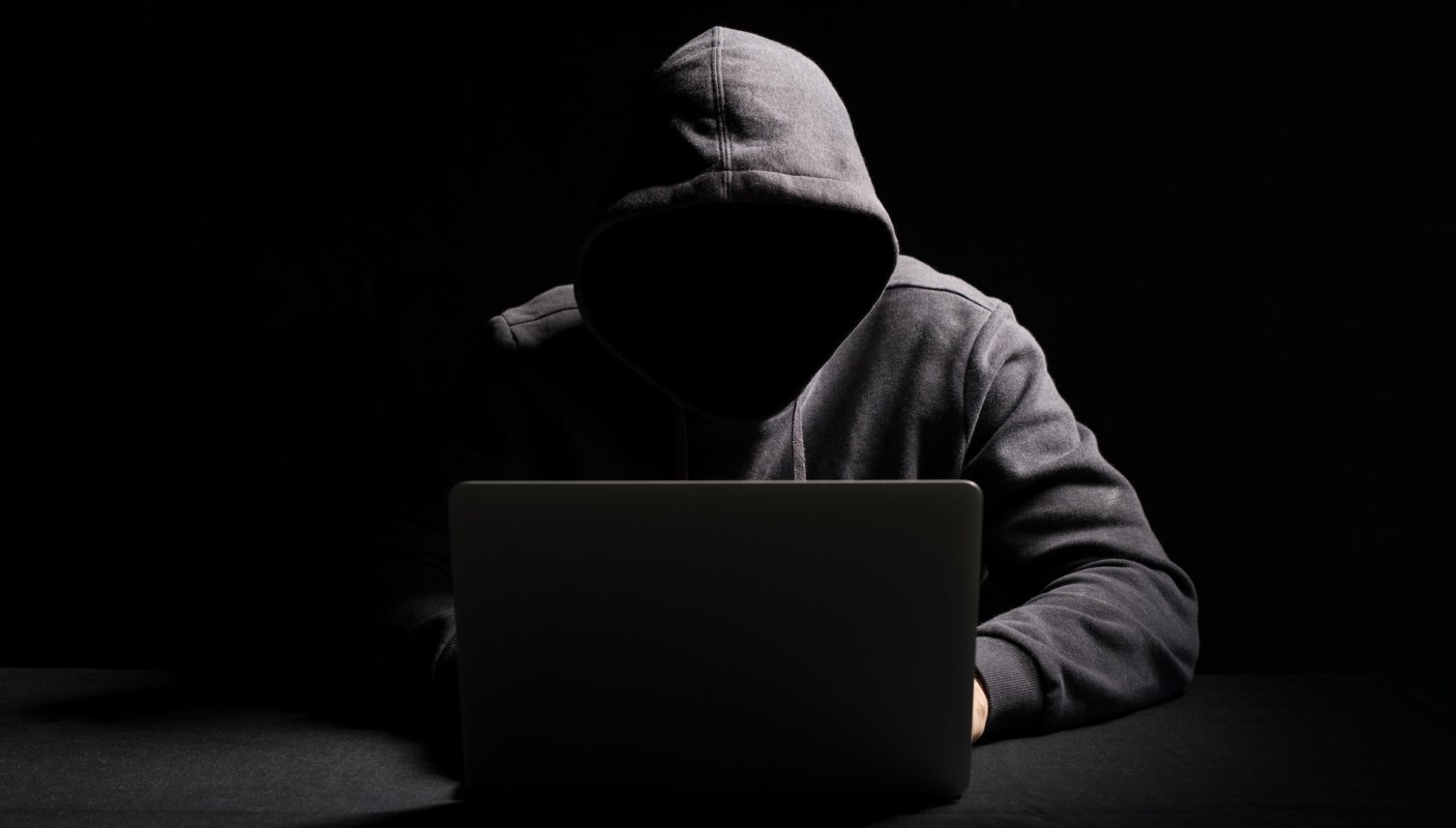 A shadowy figure in a hoodie sits in front of a laptop against a dark background.