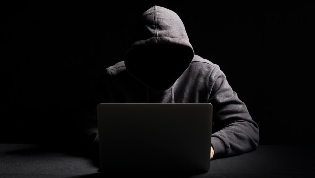 A shadowy figure in a hoodie sits in front of a laptop against a dark background.