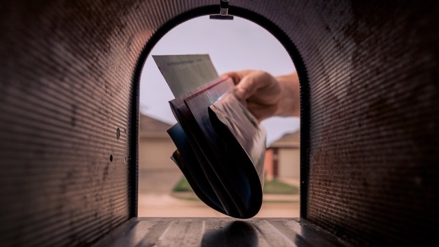 A hand is taking several letters and magazines out of a mailbox, viewed from the inside.