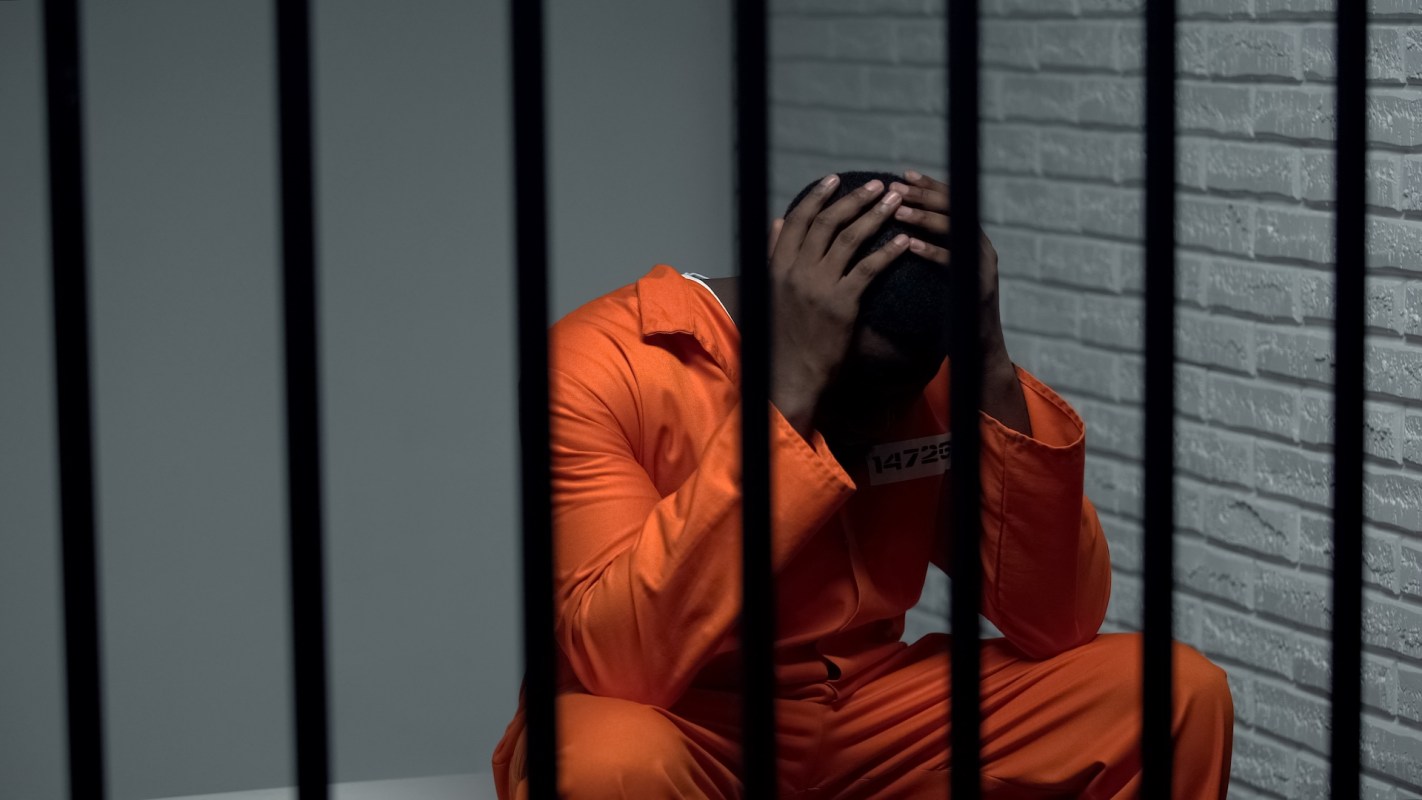 A person in an orange jumpsuit sits behind bars, holding their head in despair in a dimly lit cell.