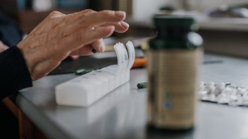 A close-up of a hand organizing pills into a weekly dose container on a table.