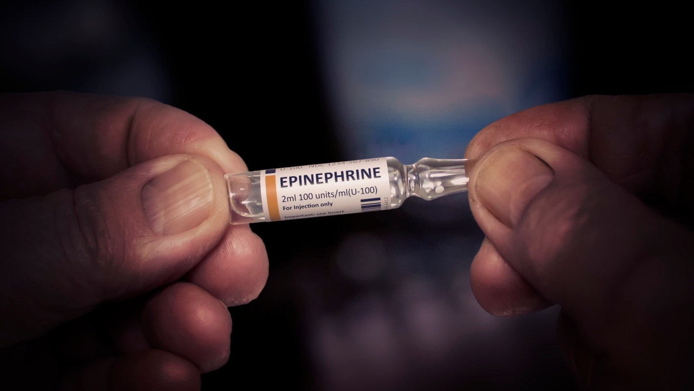 A close-up of hands holding a glass vial of epinephrine for injection.