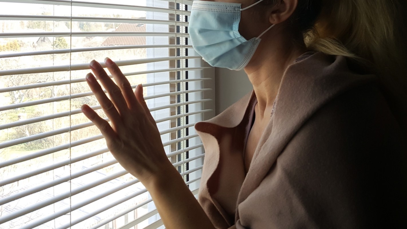 A woman wearing a medical mask looking outside wistfully through a set of blinds.