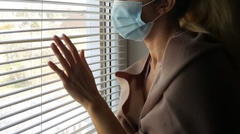 A woman wearing a medical mask looking outside wistfully through a set of blinds.