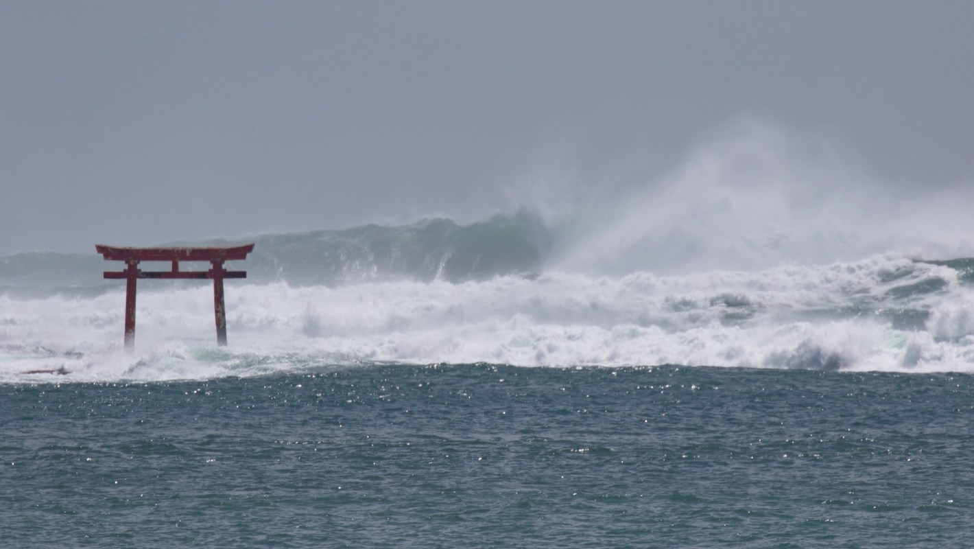 A traditional red Japanese torii gate stands in turbulent ocean waves under a gray sky.