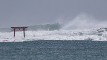 A traditional red Japanese torii gate stands in turbulent ocean waves under a gray sky.