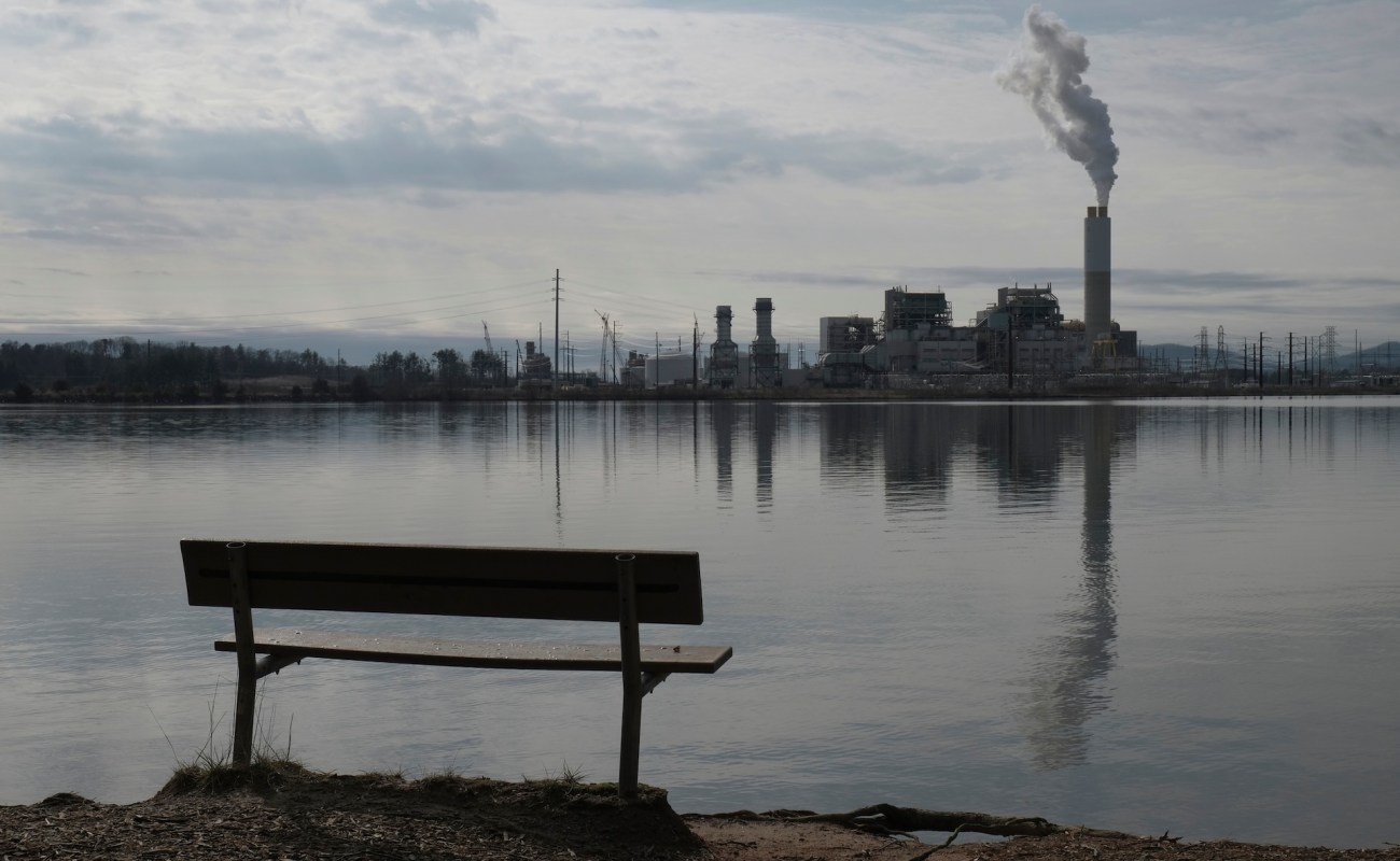 A bench by a calm lake overlooks a nearby industrial facility with smoke rising from its chimney.
