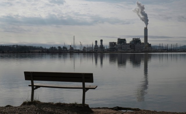 A bench by a calm lake overlooks a nearby industrial facility with smoke rising from its chimney.