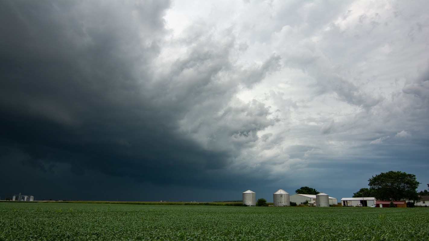 A stormy sky looms over a green farmland with silos and a white barn in the distance.