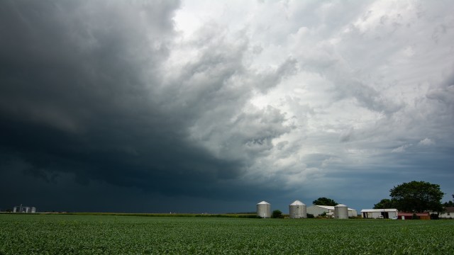 A stormy sky looms over a green farmland with silos and a white barn in the distance.