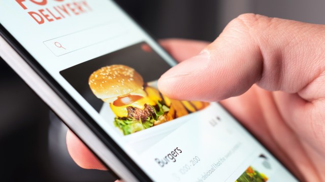 A hand holds a smartphone and scrolls through a food delivery app featuring burgers.
