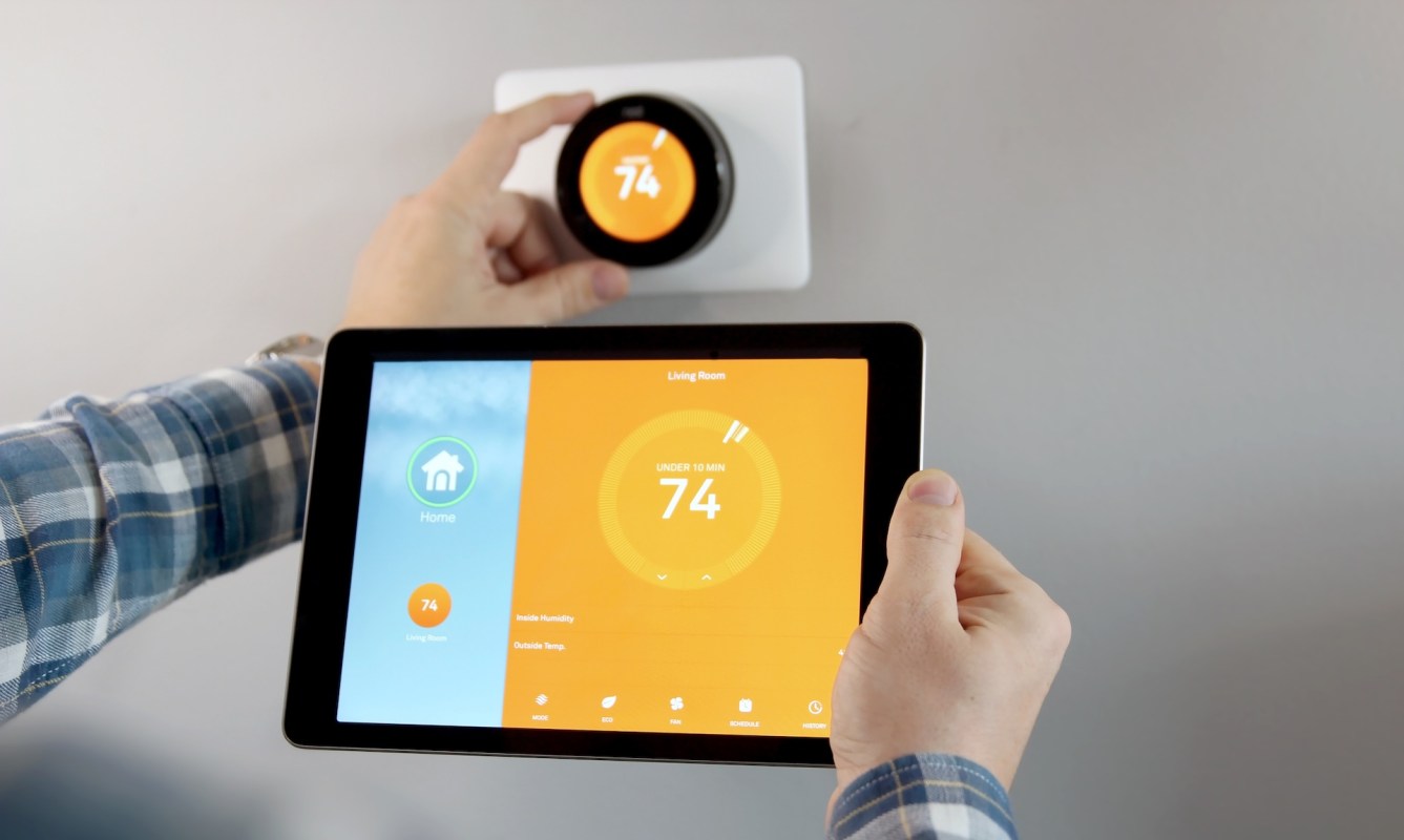 A person adjusts a smart thermostat on a wall while viewing settings on a tablet.