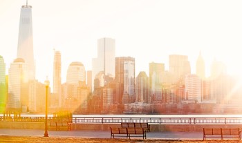 A sunlit view of a waterfront walkway overlooking a skyline with skyscrapers.