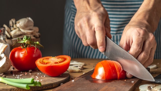 A person slices a red tomato on a wooden cutting board with green onions and mushrooms.