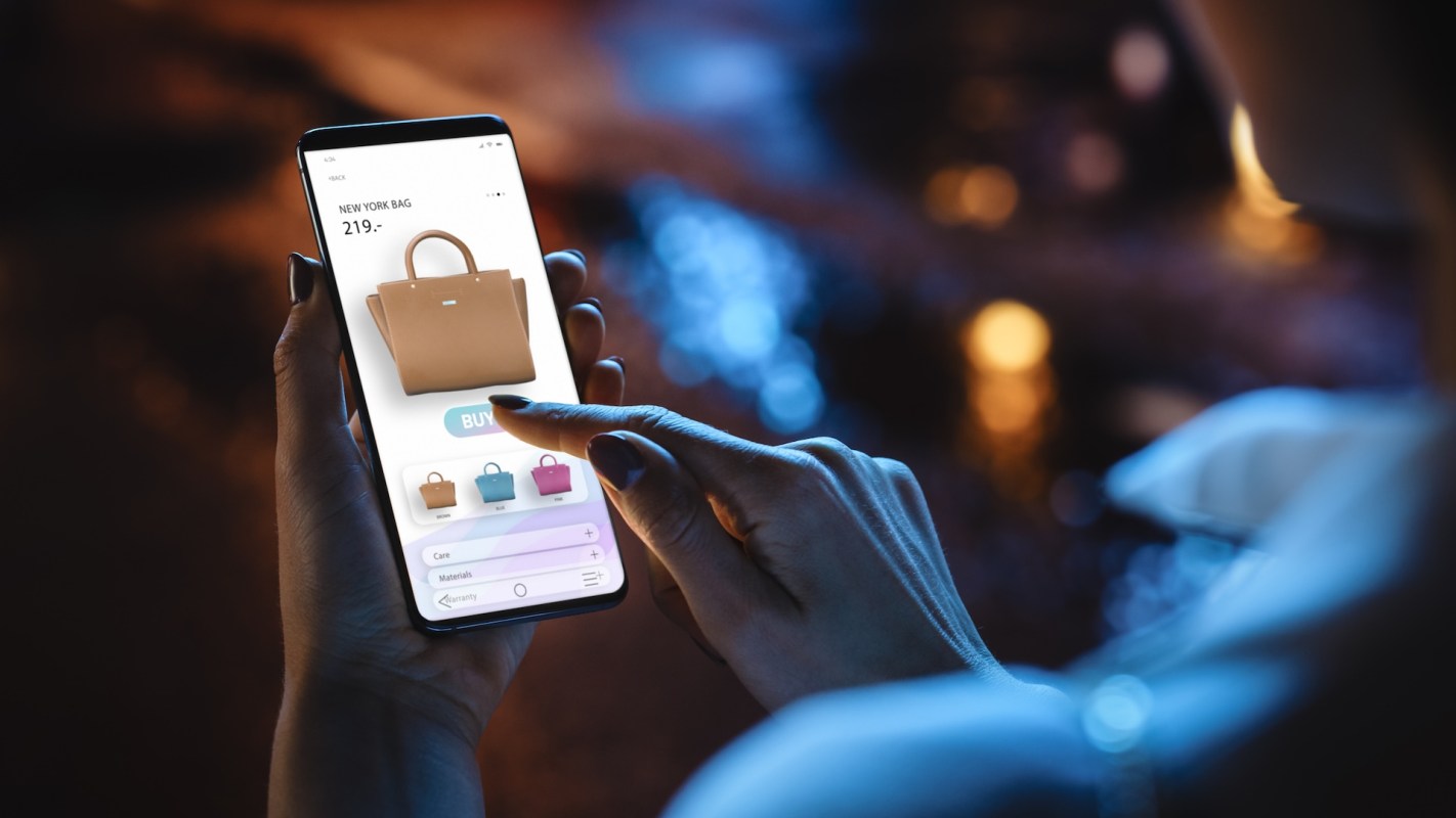 A hand holds a smartphone displaying a shopping app with a handbag and purchase options.