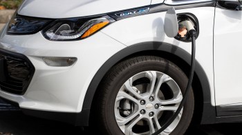 A white Chevrolet Bolt electric vehicle plugged into a charging station with a close-up of its wheel and charging port.