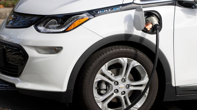 A white Chevrolet Bolt electric vehicle plugged into a charging station with a close-up of its wheel and charging port.