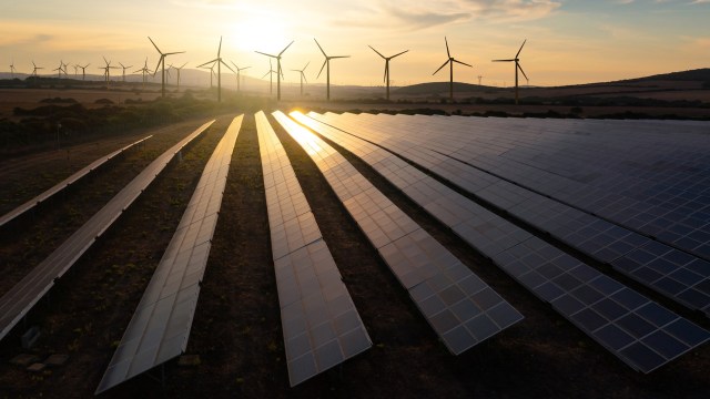 Aerial view of solar panels with wind turbines in the background at sunset.