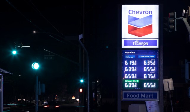A brightly lit Chevron gas station sign displaying fuel prices at night, with a green traffic light in the background.