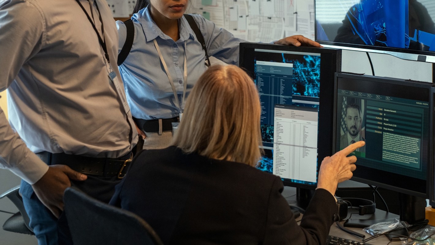Three people examine computer screens displaying data and an image of a man in a modern office setting.