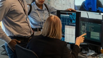 Three people examine computer screens displaying data and an image of a man in a modern office setting.