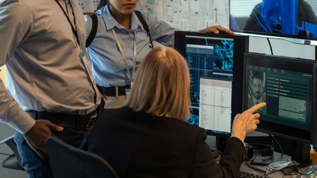 Three people examine computer screens displaying data and an image of a man in a modern office setting.