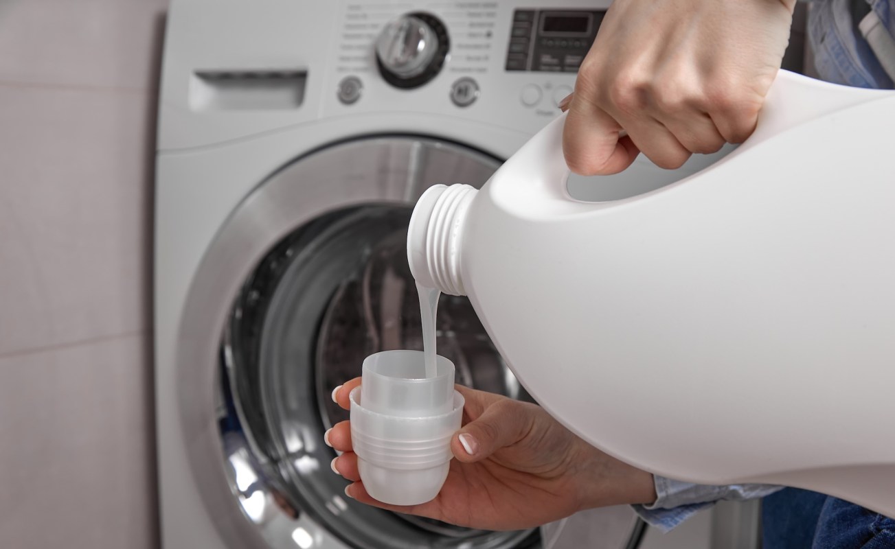 A person pours liquid detergent from a large bottle into a measuring cup near a washing machine.