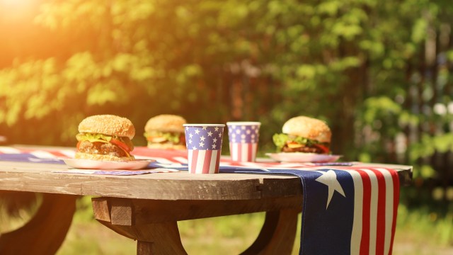 A picnic table set with burgers and cups featuring the American flag, with bright sun coming from the upper left.