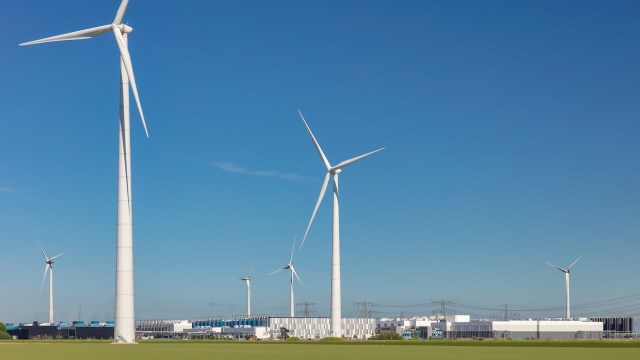 A row of large wind turbines against a clear blue sky, with an industrial building in the background.
