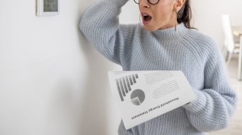 A woman in a blue sweater looks shocked while holding an energy usage statement with graphs next to a thermostat.