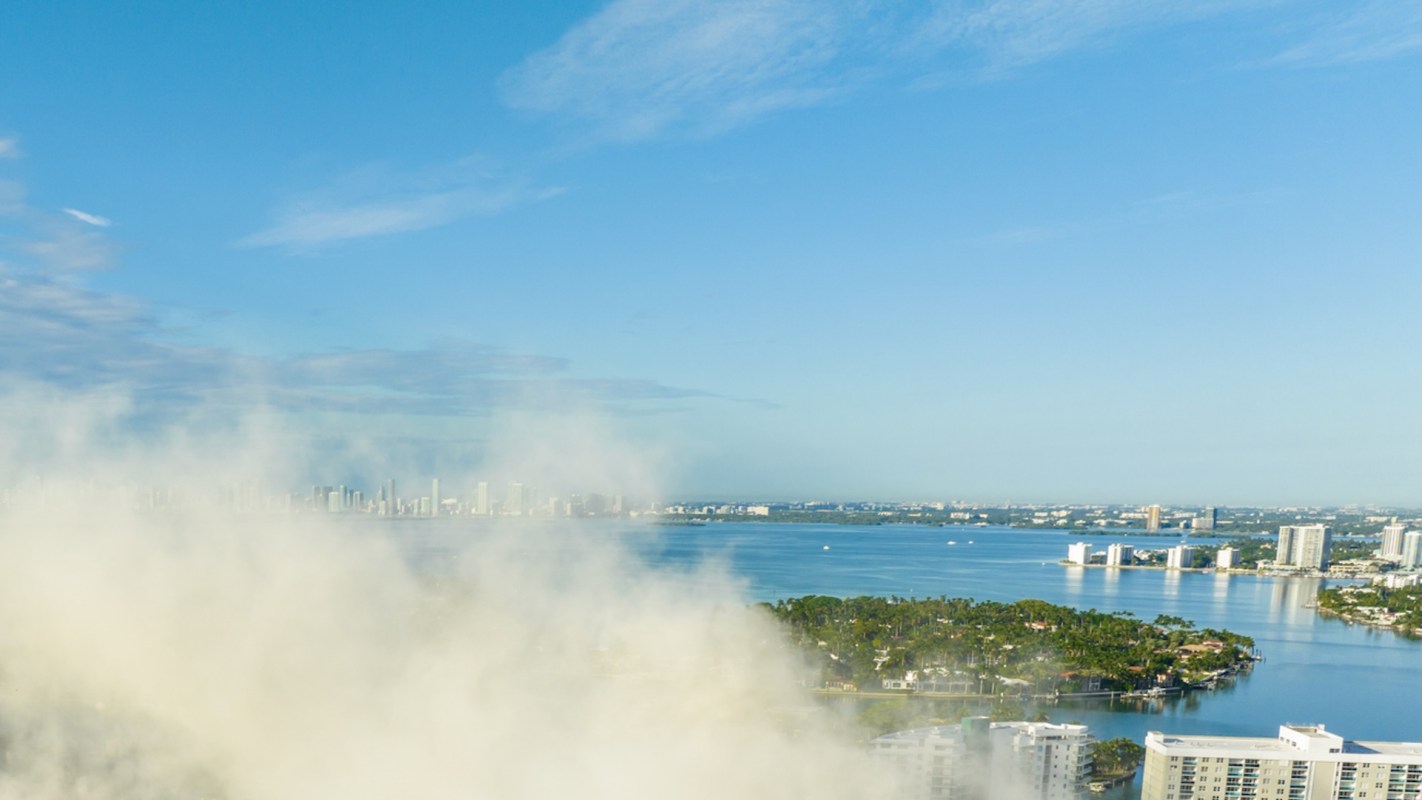 A panoramic view of a city skyline across a bay under a clear blue sky with smoke in the foreground.