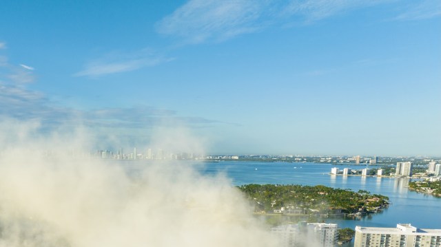 A panoramic view of a city skyline across a bay under a clear blue sky with smoke in the foreground.