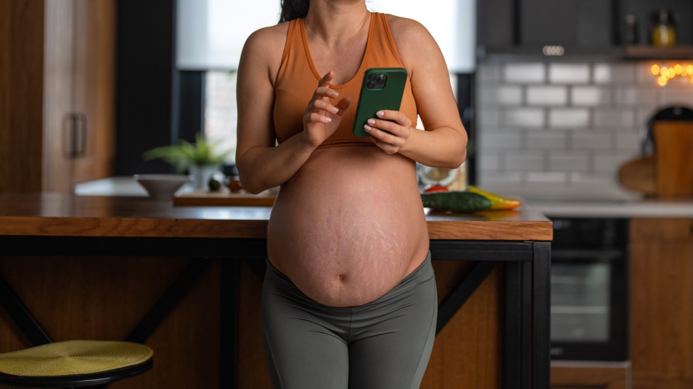A pregnant person in a sports top checks their phone while standing in a kitchen.