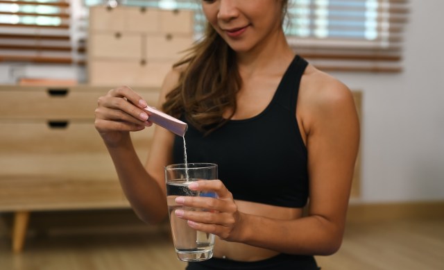 A woman in a black sports bra adds a powdered collagen supplement to a glass of water.