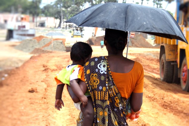 A woman carrying a child under an umbrella on a hot day.
