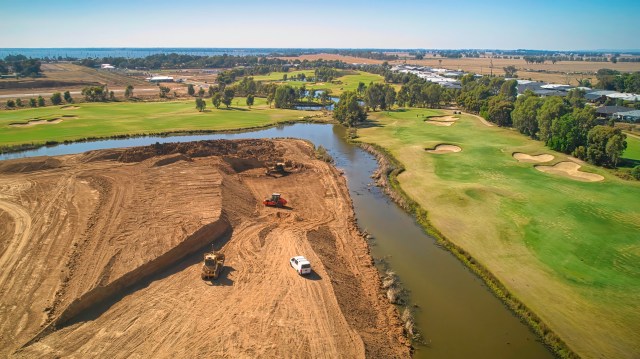An aerial view of a construction site near a golf course, with heavy machinery and a winding river.