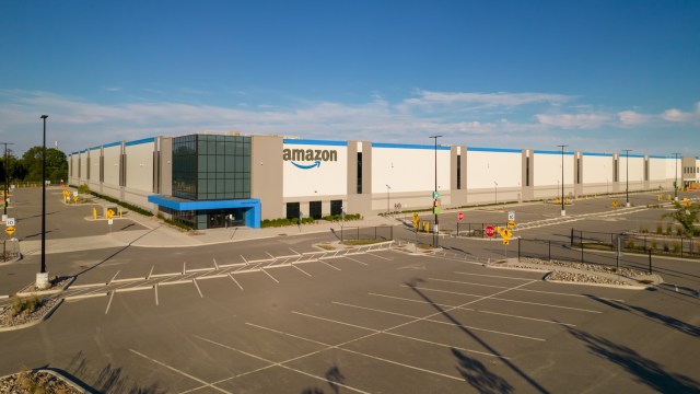 A large Amazon distribution warehouse with a blue and gray exterior, surrounded by an empty parking lot.