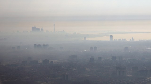 A hazy skyline of Toronto with a prominent tower, enveloped in fog and smog, against a muted sky.