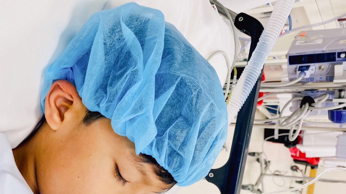 A child with a blue surgical cap resting on a hospital bed surrounded by medical equipment.