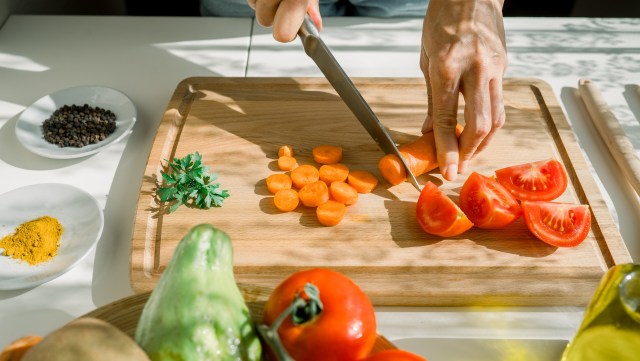 A person slices tomatoes and carrots on a wooden cutting board surrounded by fresh herbs and spices.