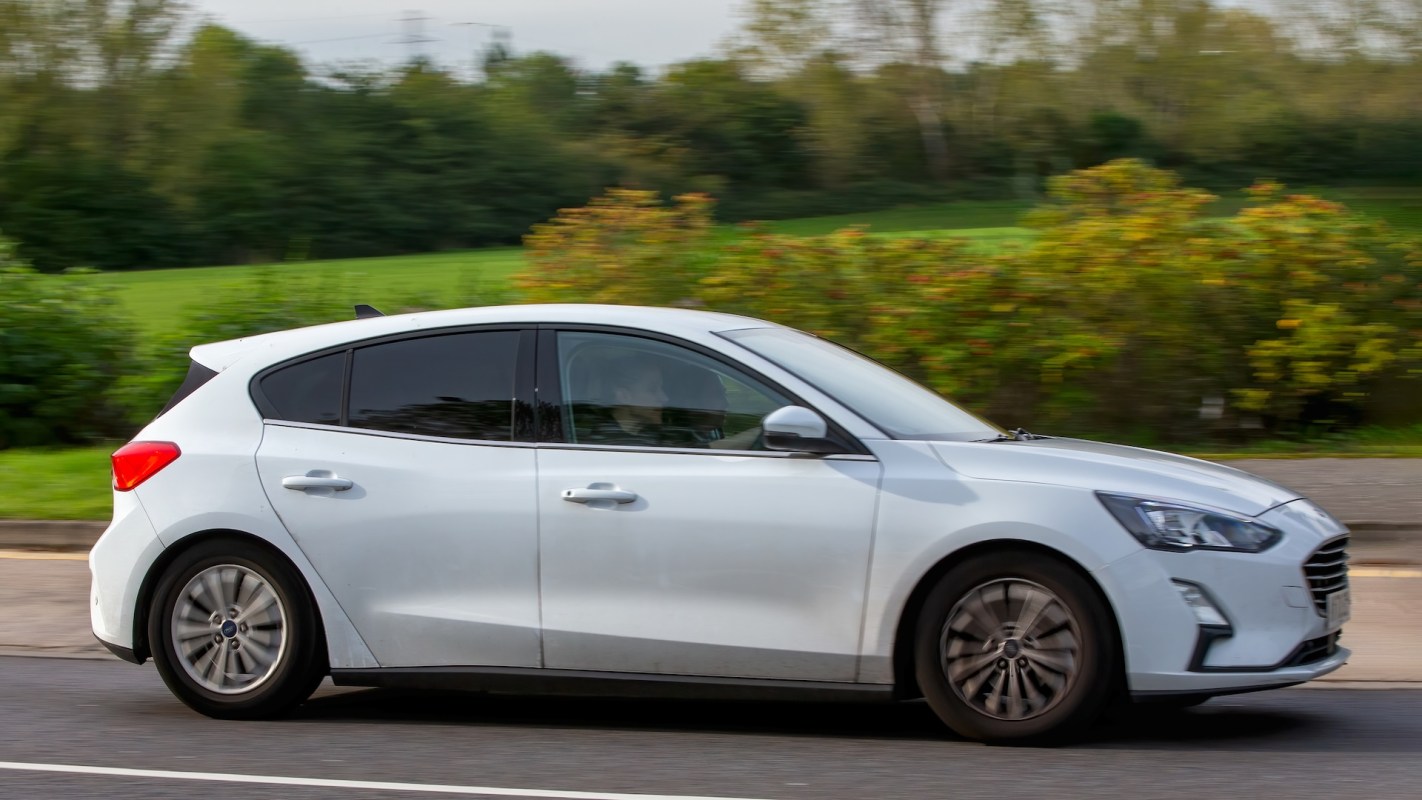 A silver hatchback car drives past greenery on the side of a road.