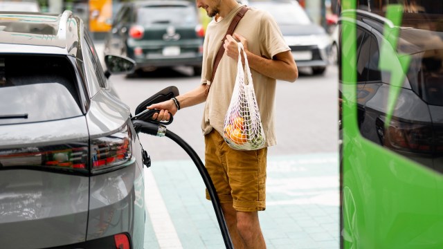 A man charges a gray electric car while holding a mesh bag of groceries in a service station parking lot.