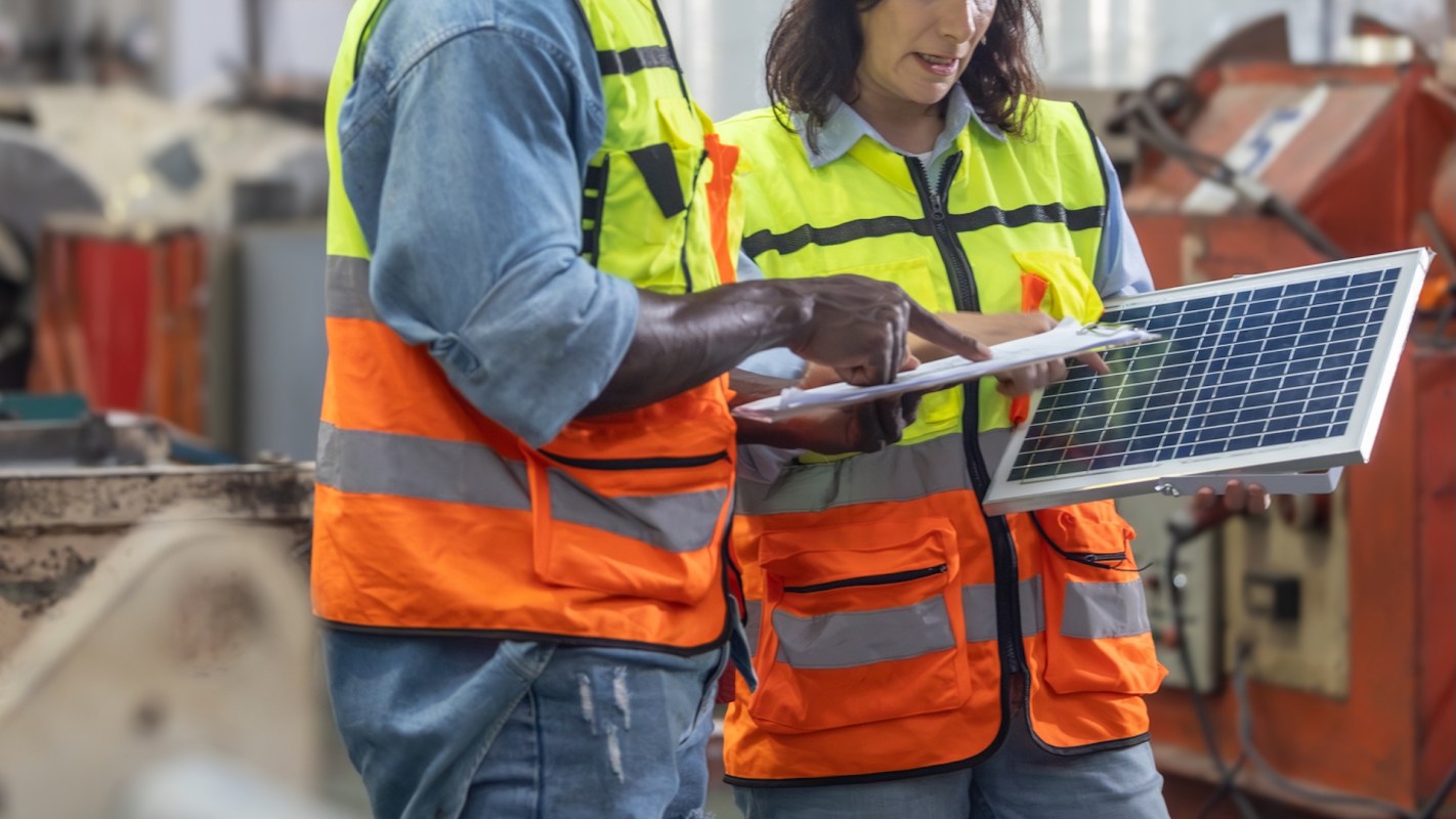 Two workers in safety vests discuss a solar panel while reviewing documents in an industrial setting.