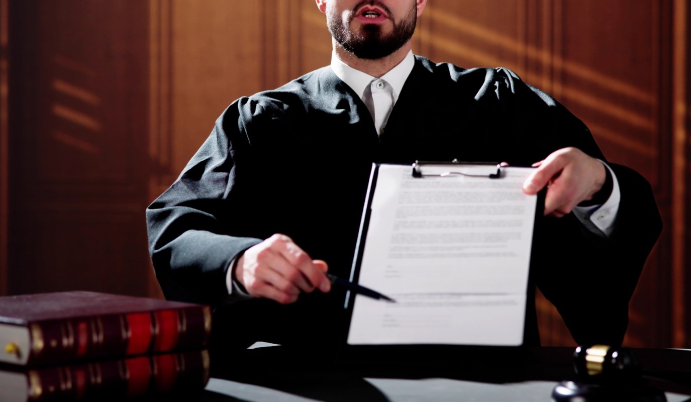 A judge in black robes angrily holds a clipboard with a document and points at it, seated at a desk in a courtroom.