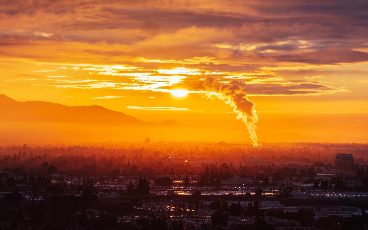 A sunset casts an orange glow over a cityscape with smoke rising from industrial buildings.