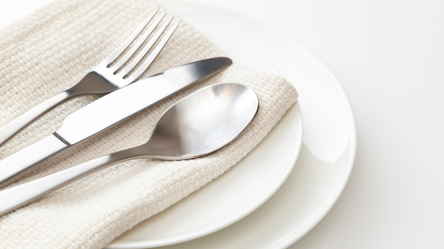 A plate set with a knife, fork, and spoon resting on a beige cloth napkin.