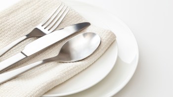 A plate set with a knife, fork, and spoon resting on a beige cloth napkin.