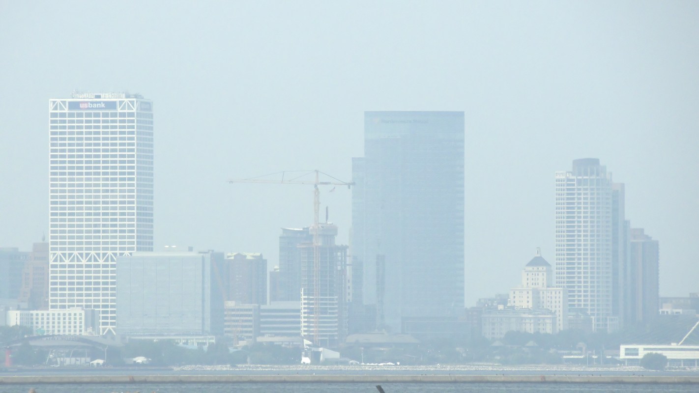 A skyline view of Milwaukee with tall buildings and a construction crane, shrouded in haze.