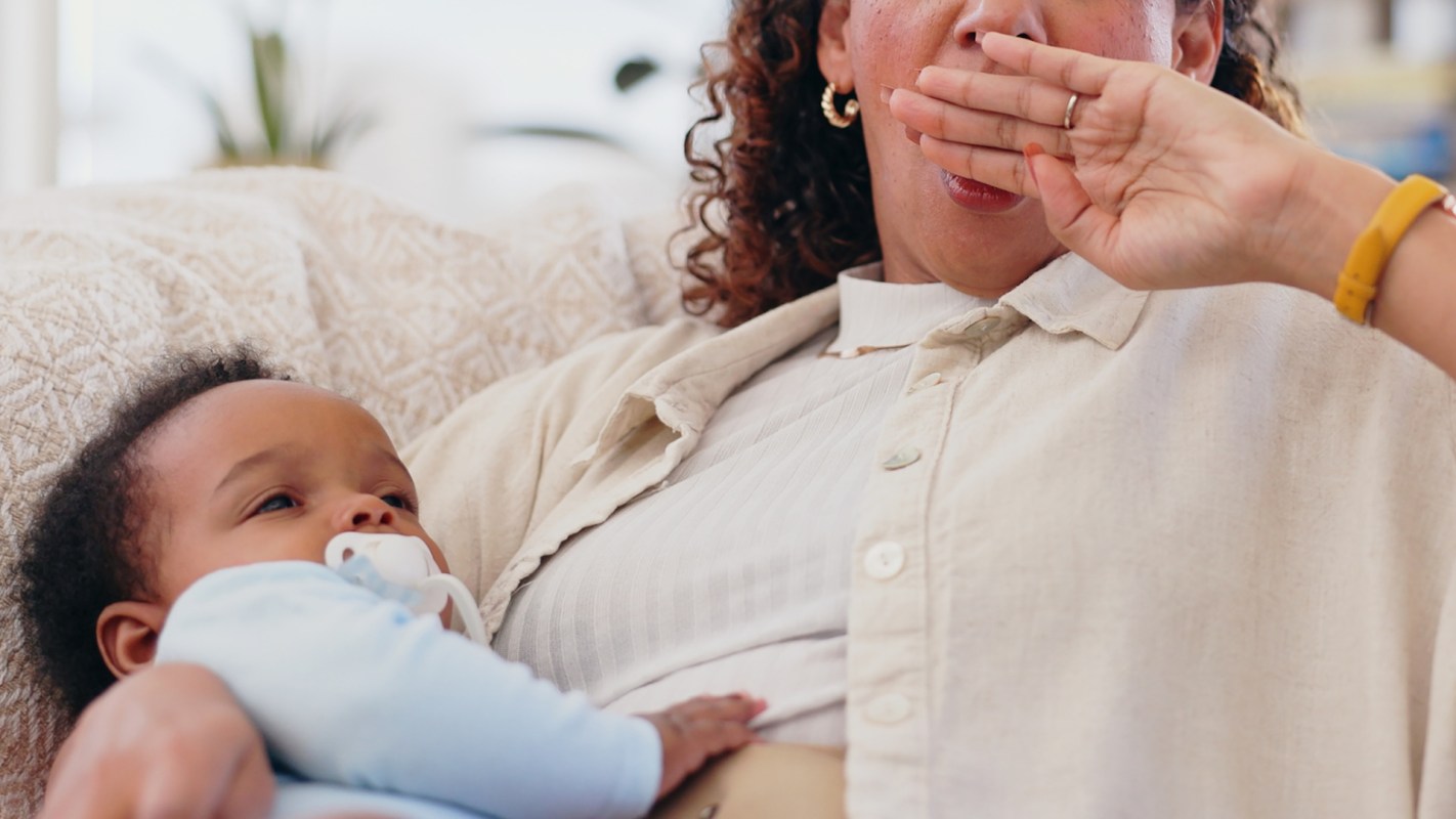 A tired parent yawns while holding a baby who is sucking on a pacifier on a cozy couch.
