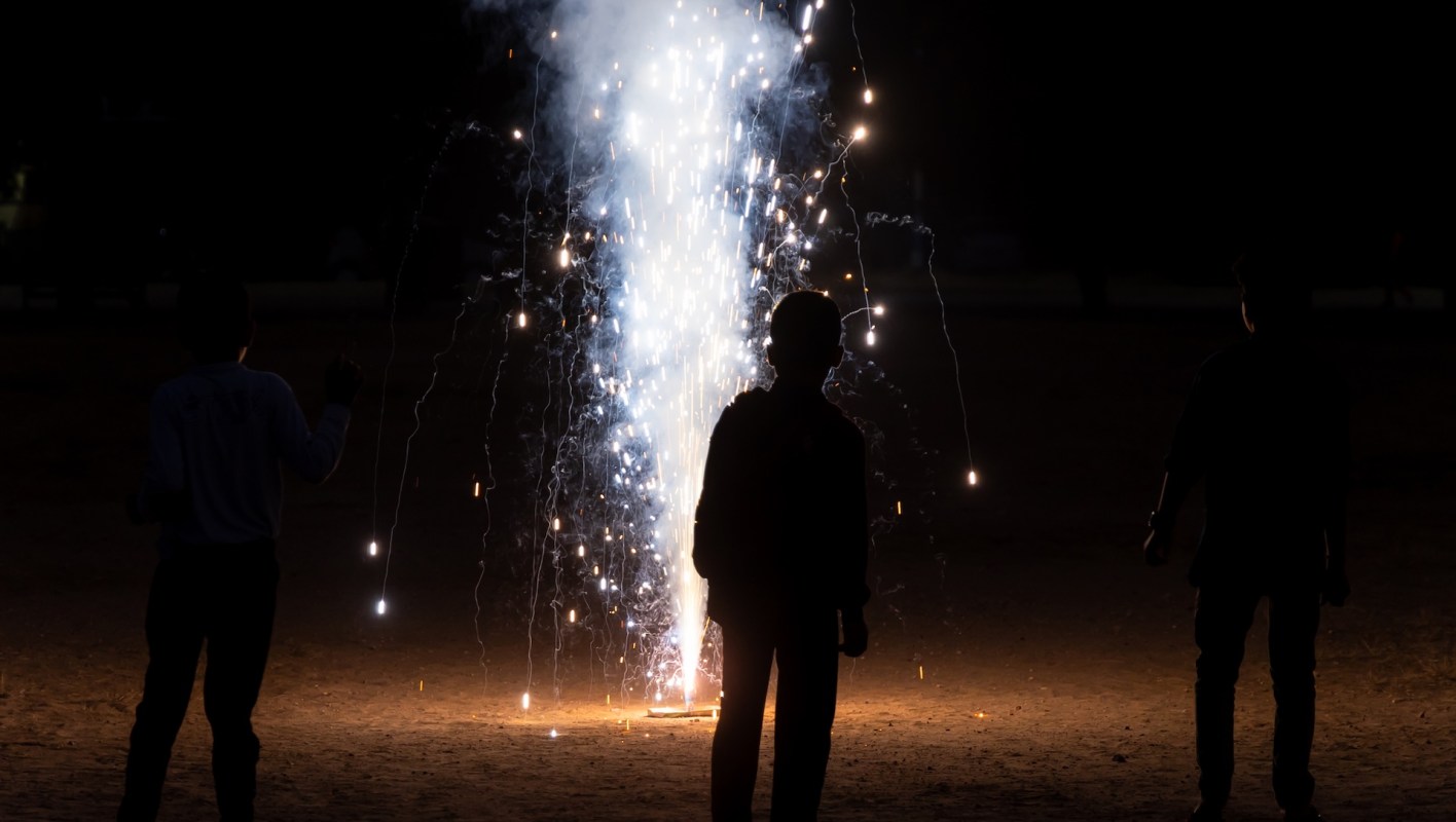 Three children silhouetted against a bright fountain firework illuminating a dark outdoor setting.